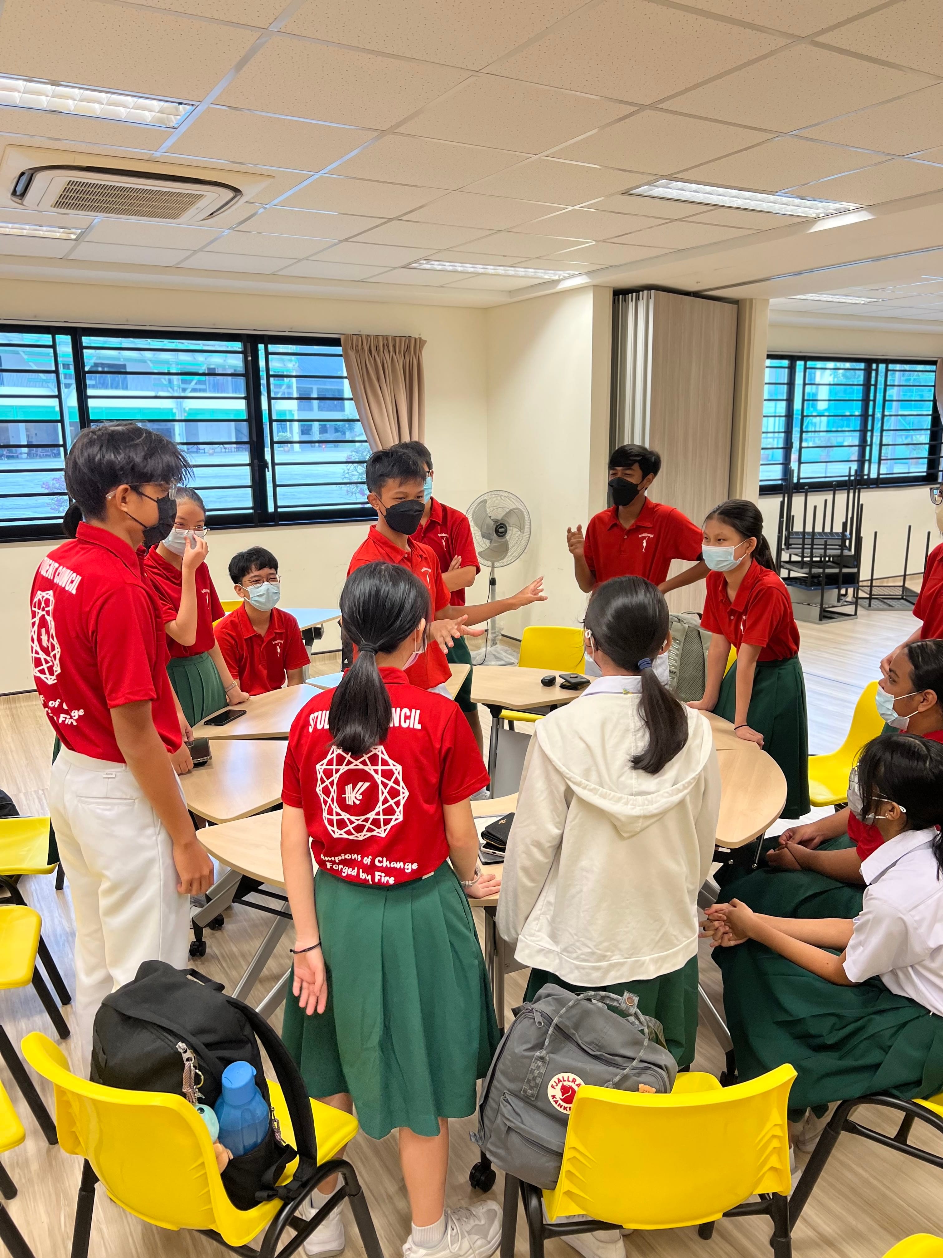 Students in red Student Council shirts and green skirts stand and sit in a circle discussing in a classroom.